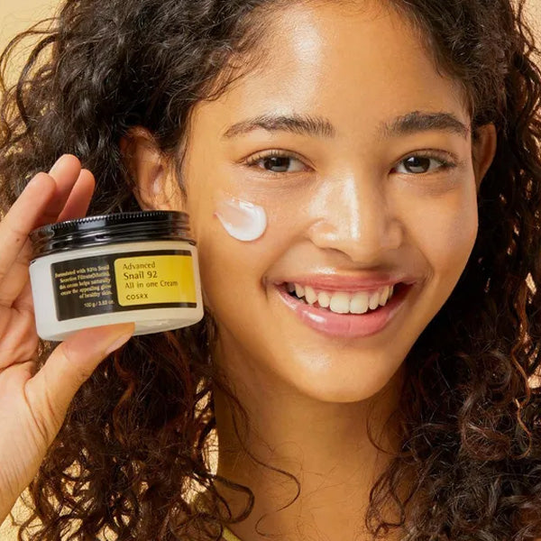 Woman smiling with a jar of COSRX Advanced Snail 92 All In One Cream, applying cream to face