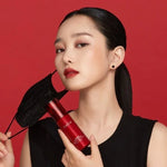 Woman with sleek hair holding TIRTIR makeup fixer spray and a black face mask, red background